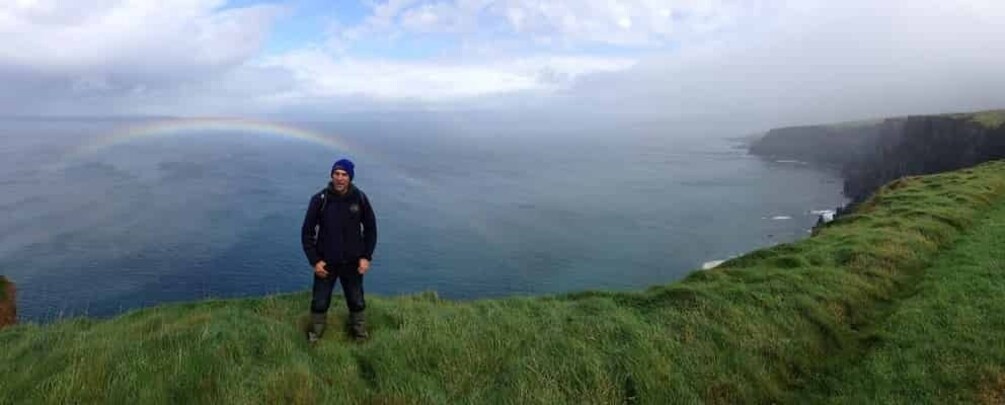 A person standing on a grassy cliff overlooking a vast body of water with a rainbow in the distance. 5