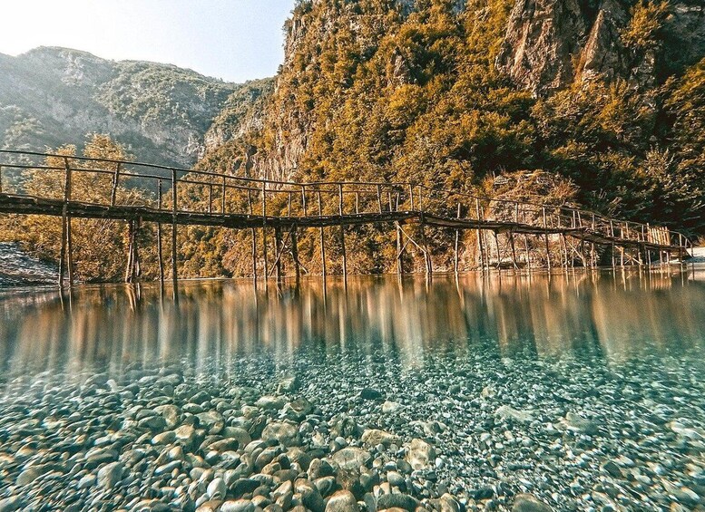 A wooden bridge over a clear lake with rocky shore, surrounded by autumn-colored trees and mountains. 4
