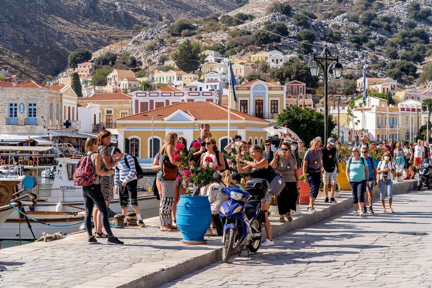 A crowded waterfront promenade with people walking, a blue motorcycle parked, and traditional buildings in the background. 2