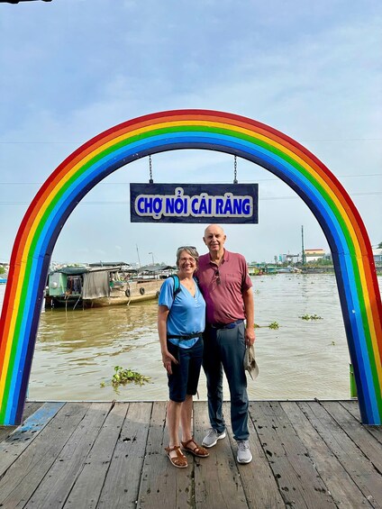 Two people standing under a rainbow-colored arch with a sign that reads "CHO NOI CAI RANG". 3