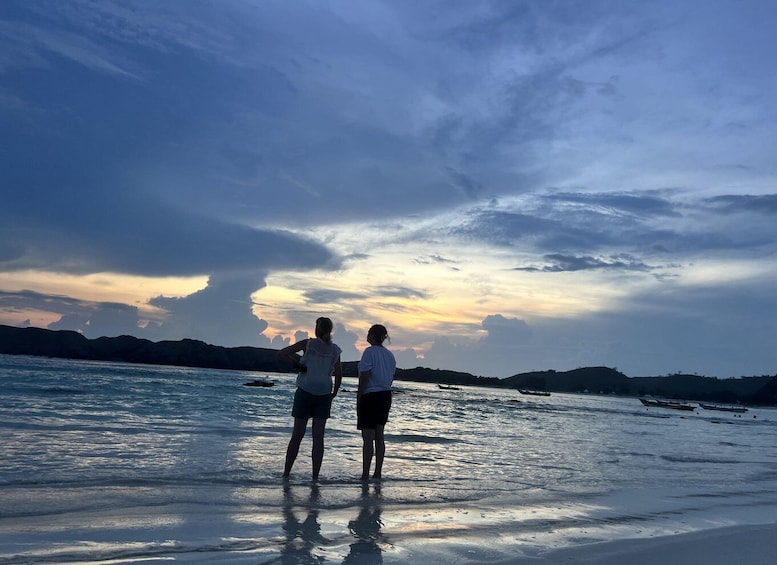 Two people standing on a beach at sunset with a calm sea and distant hills. 3