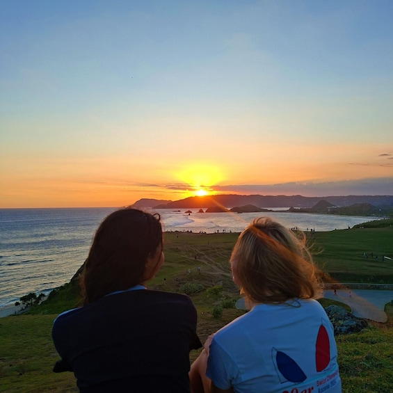 Two people overlooking a sunset at a coastal area with grassy terrain and distant hills. 2