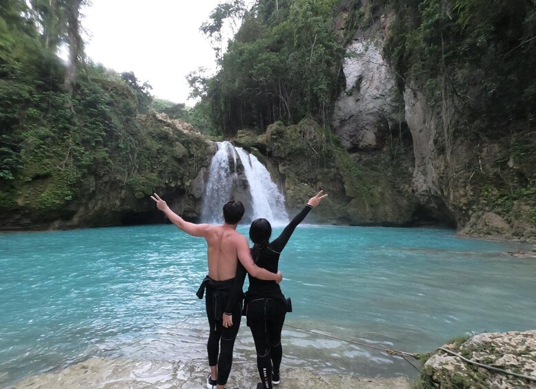 Two people standing by a turquoise river with a waterfall in the background. 1
