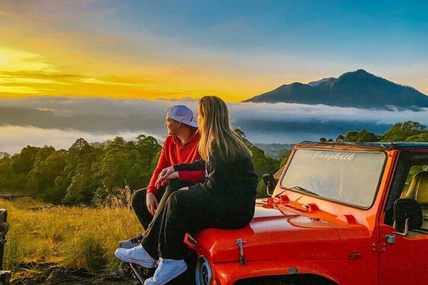 Two people sitting on a red vehicle with a scenic view of a mountain and clouds in the background. 1