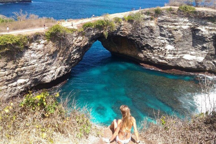A natural rock arch over a clear blue pool with a person sitting at the edge. 4