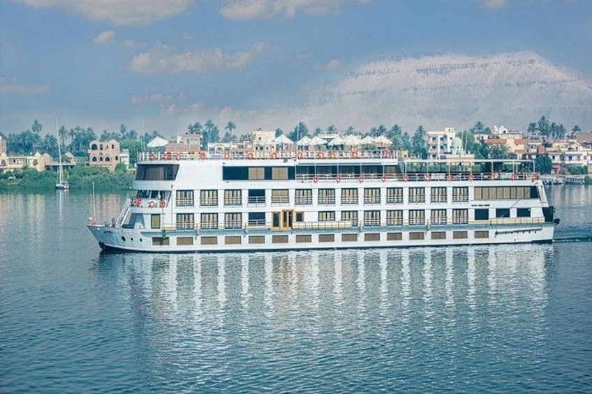 A large riverboat with multiple decks, floating on calm water, with a cityscape and mountains in the background. 1