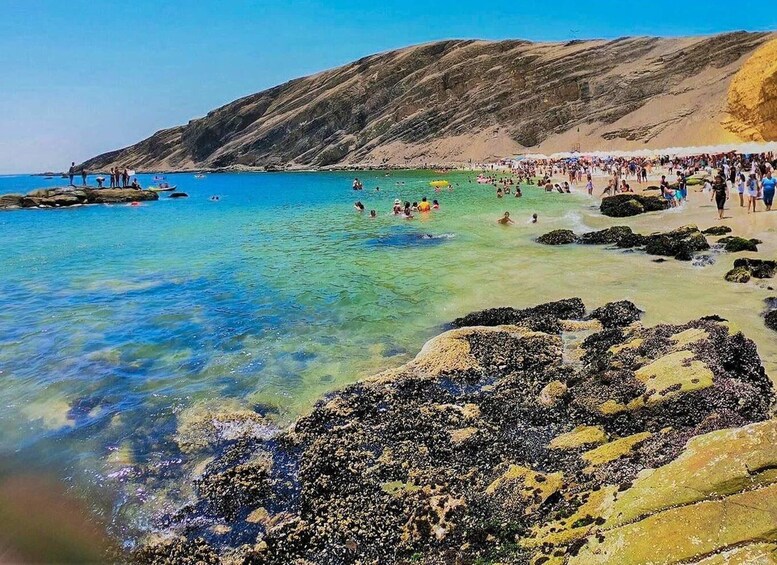 Ein Strand mit klarem türkisblauem Wasser, felsigem Ufer und einer Menschenmenge. 5