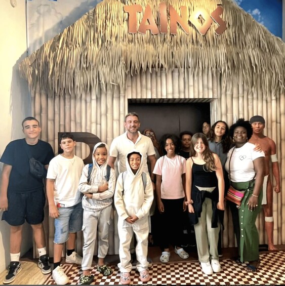 A group of people posing in front of a thatched-roof hut with the word "TAKES" on it. 2
