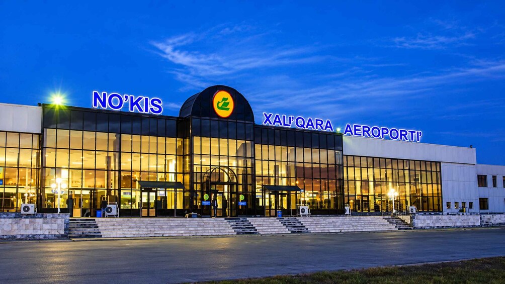 An airport terminal with illuminated signs reading 'NO'KIS' and 'XAL'QARA AEROPORT'. 3