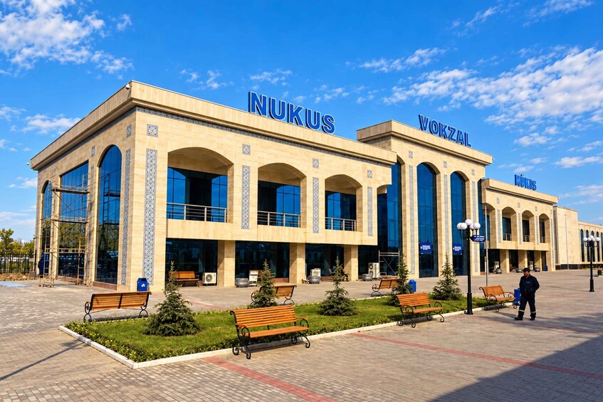 A modern building with the name 'NUKUS' and 'VOKZAL' on the facade, surrounded by a paved area with benches and a clear sky. 2
