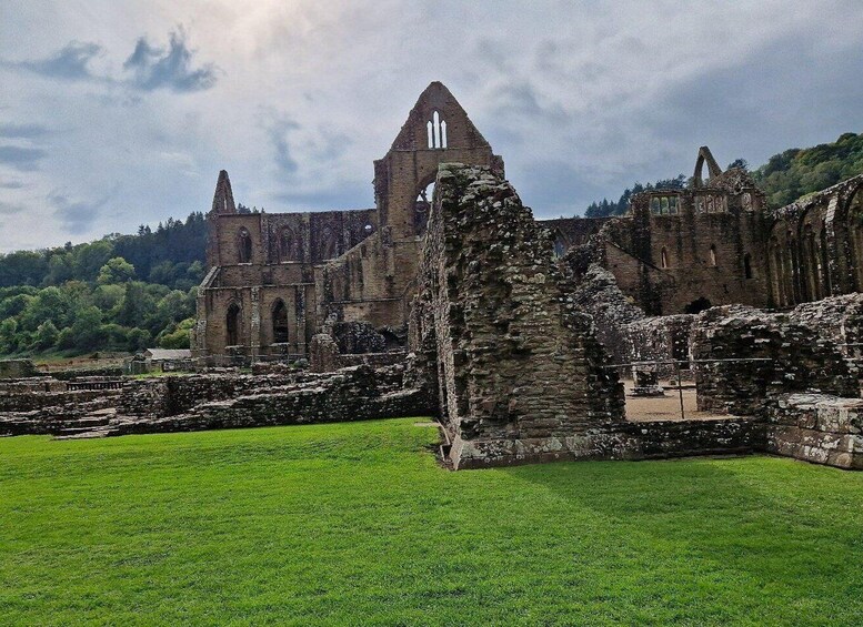 The ruins of a large, historic stone building with multiple arches and towers, surrounded by lush green grass and trees. 3