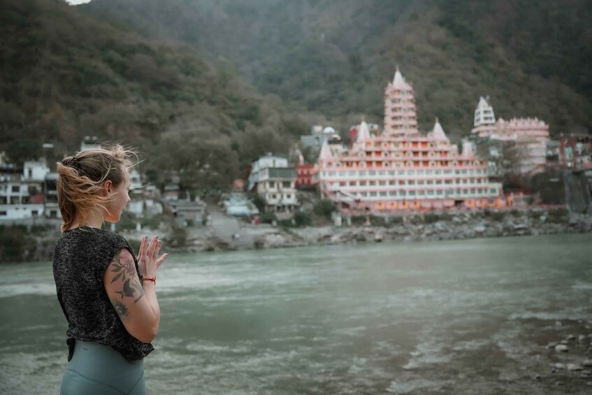 A person with tattoos praying by a river with a temple in the background. 5