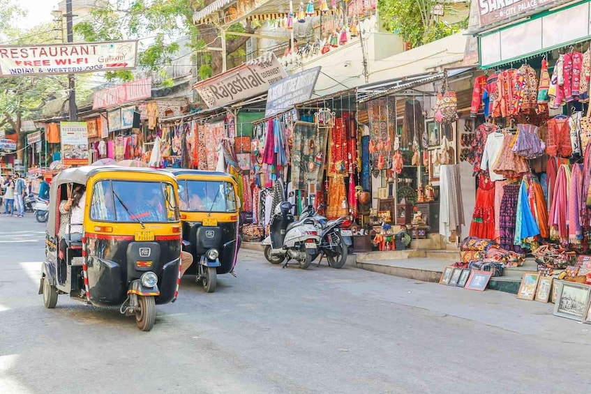 A busy street with auto-rickshaws, shops selling colorful textiles, and a sign for a restaurant. 1