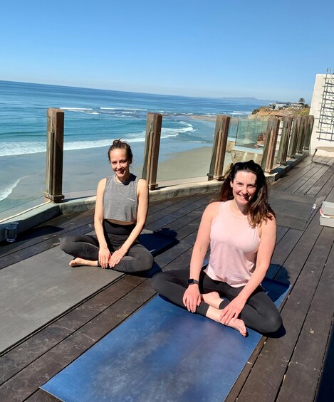 Two people sitting on yoga mats on a wooden deck by the ocean. 1