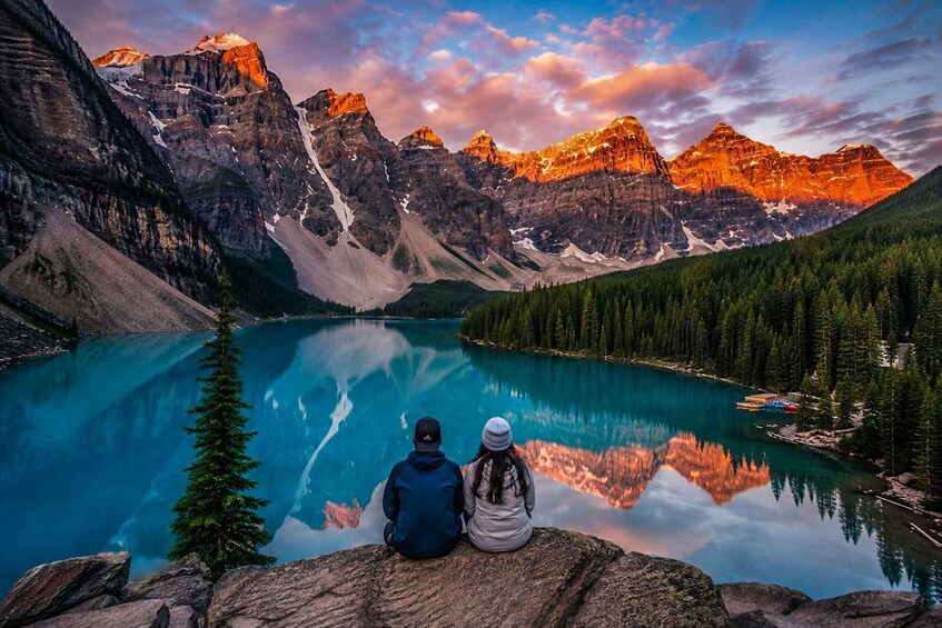 Two people sitting on a rock overlooking a turquoise lake with mountains in the background. 1