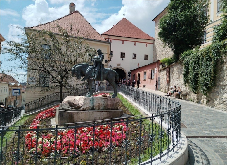 A statue of a man on horseback in a public square with red flowers and a stone building in the background. 2