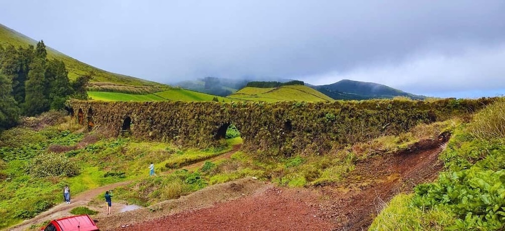 A stone bridge over a muddy path in a hilly landscape with green fields and a cloudy sky. 3