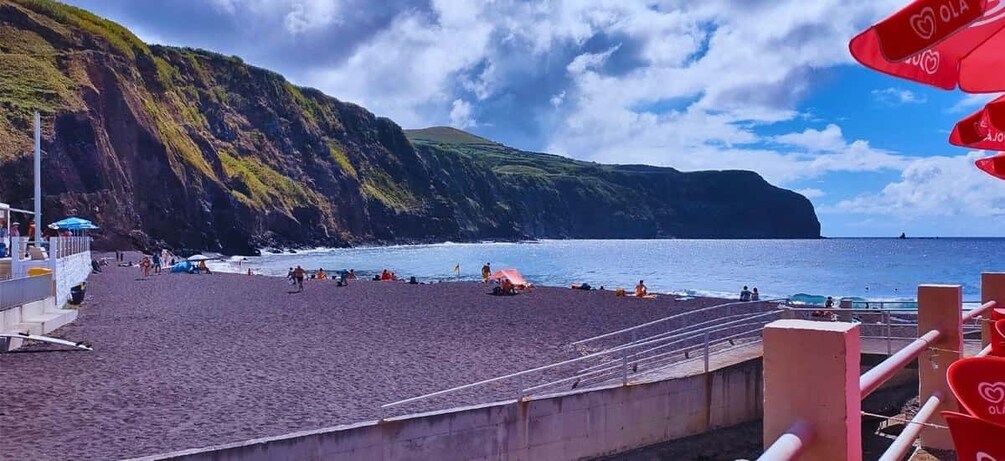 A beach with people sunbathing and swimming, a rocky cliff, and a clear sky. 4
