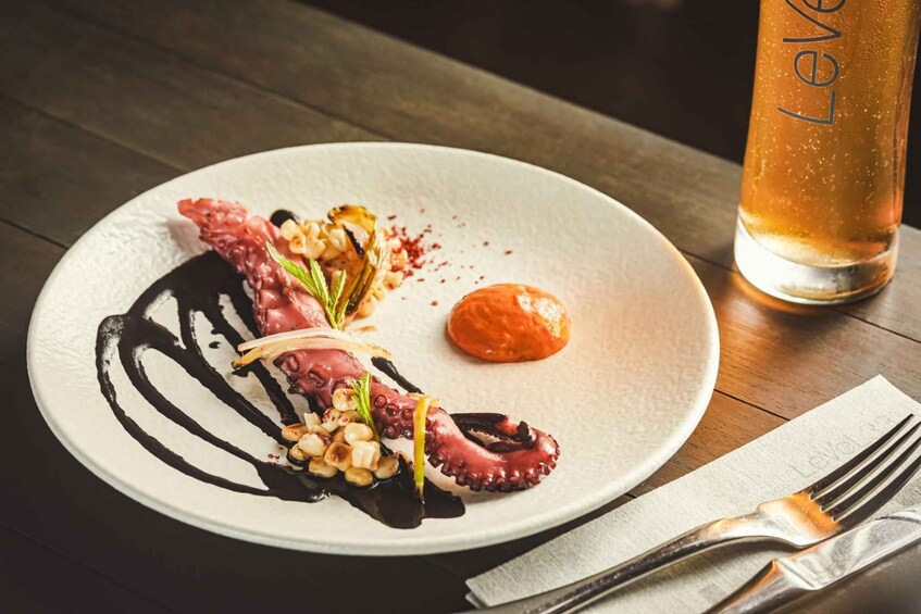 A plate of octopus dish with sauce and garnish, a glass of beer, and a fork on a wooden table. 2