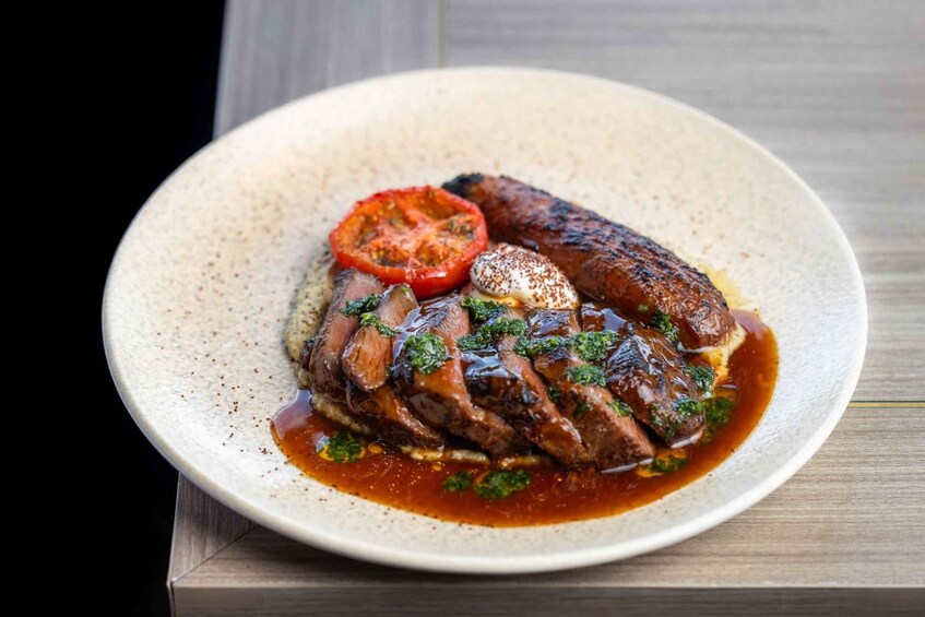 A plate of food with a grilled sausage, sliced meat, a tomato slice, and a poached egg, garnished with herbs. 3