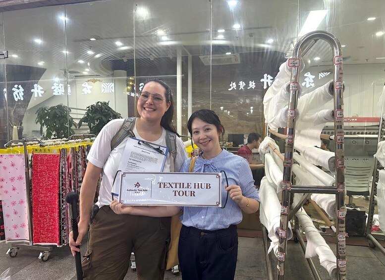 Two people holding a sign that reads 'TEXTILE HUB TOUR' in front of a store with fabric rolls and a metal rack. 5