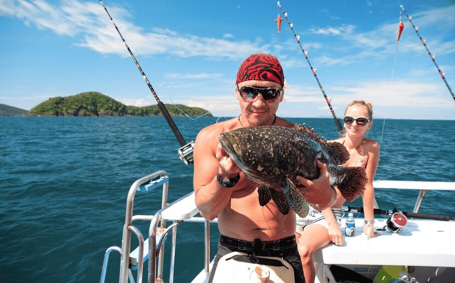 A man holding a fish on a boat with another person seated nearby, fishing rods in the background, and a small island in the distance. 4