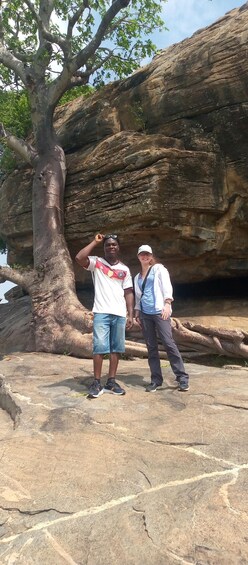 Two people standing on rocky ground with a large tree and a rock formation in the background. 1