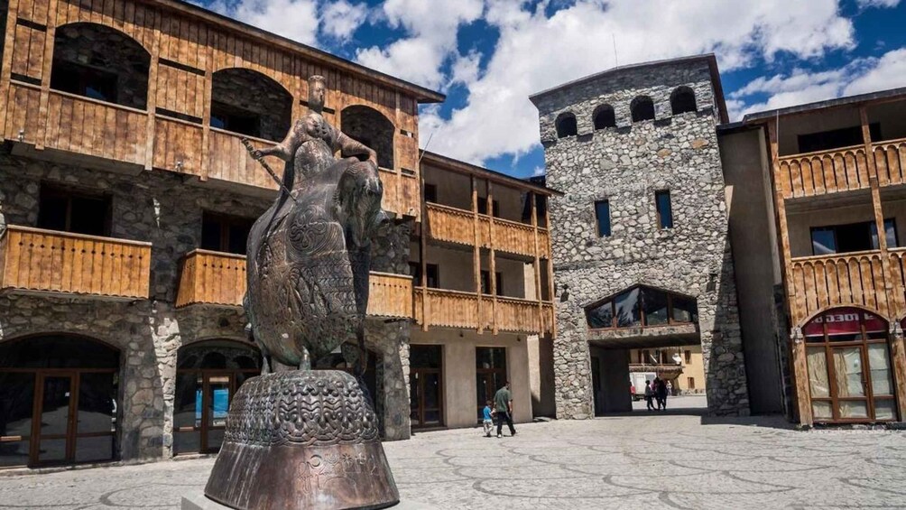 A courtyard with a large bronze statue, stone buildings with balconies, and a stone tower. 3
