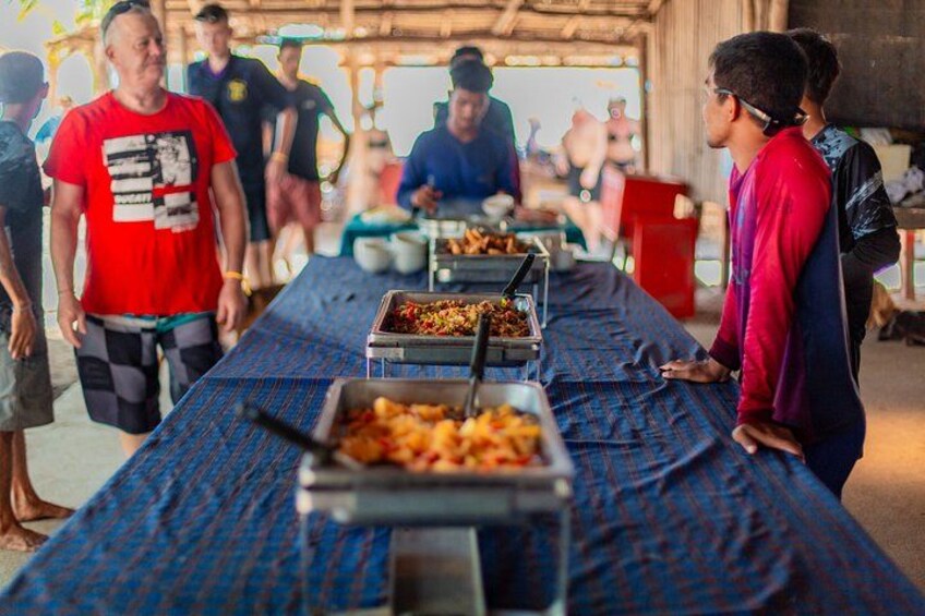 A communal dining area with a long table covered with a blue cloth, food trays, and people standing around. 5