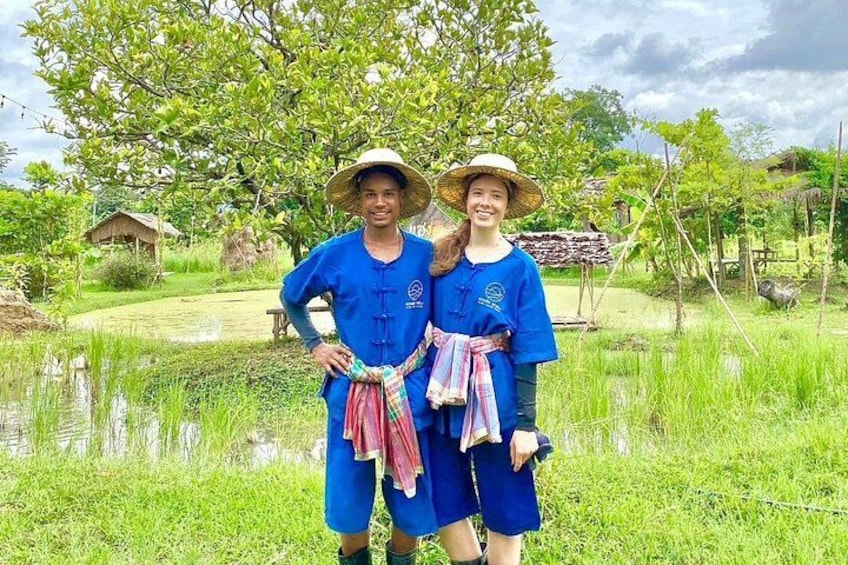 Twee personen in blauwe traditionele kleding staan in een weelderig groen veld, met een vijver en bomen op de achtergrond. 2