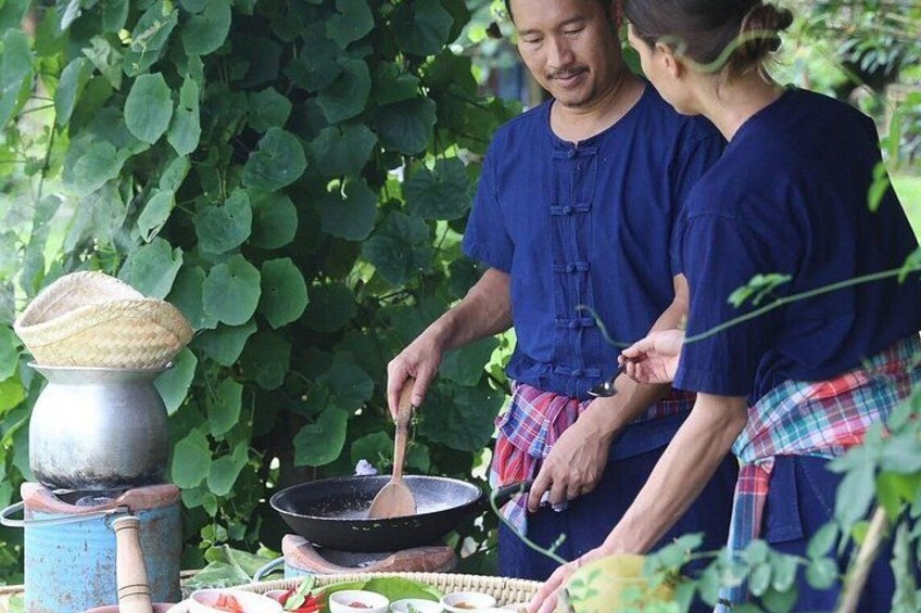 Twee personen koken buiten, met een pan op een fornuis, omgeven door groen. 4
