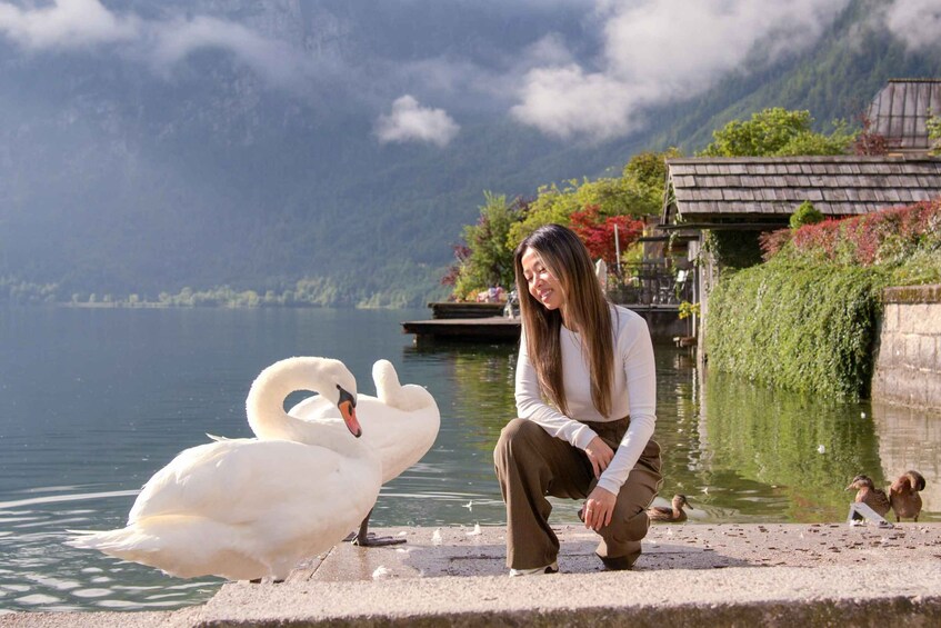 A woman sitting by a lake with swans and ducks, surrounded by greenery and mountains. 2