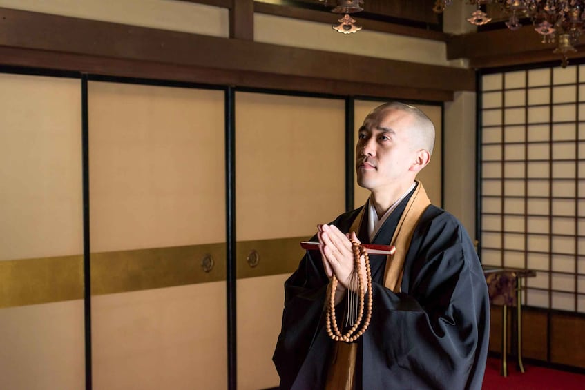 A person in traditional attire holding prayer beads in a room with sliding doors and a hanging ornament. 2