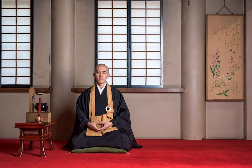 A person in traditional attire sitting on a red carpet in a room with large windows and a hanging scroll. 1