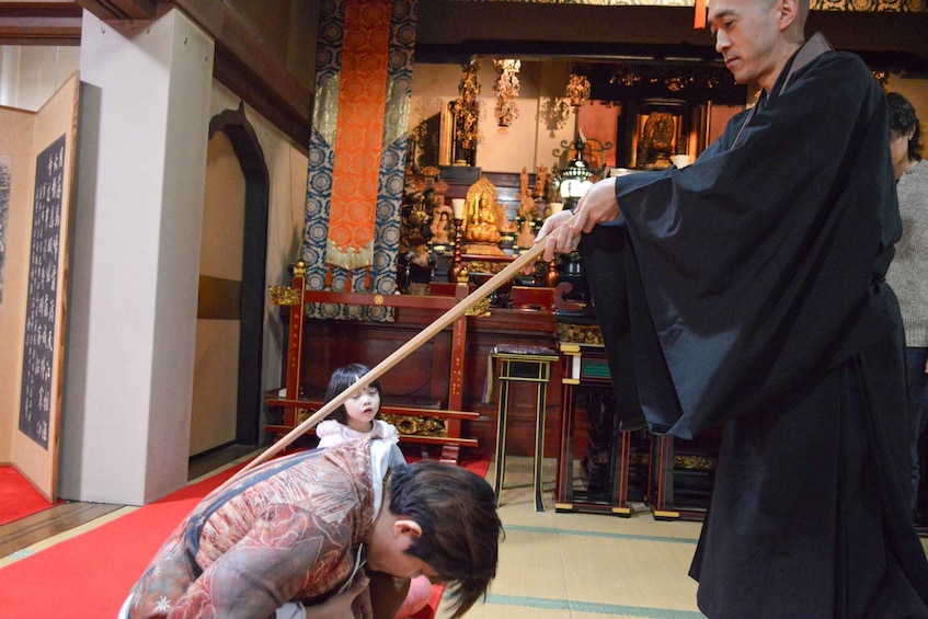 A person in traditional attire holding a long stick, possibly a ceremonial object, in a room with ornate decorations and a red carpet. 5