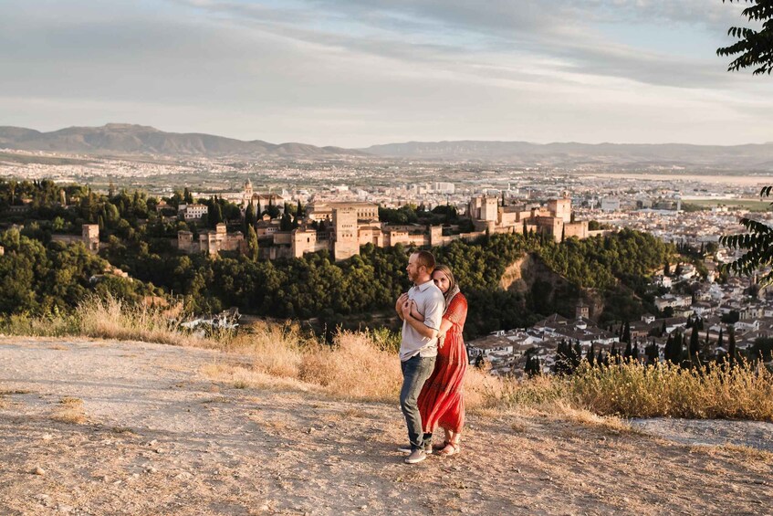 A couple embracing on a rocky outcrop overlooking a historic city with medieval architecture and a mountainous backdrop. 2