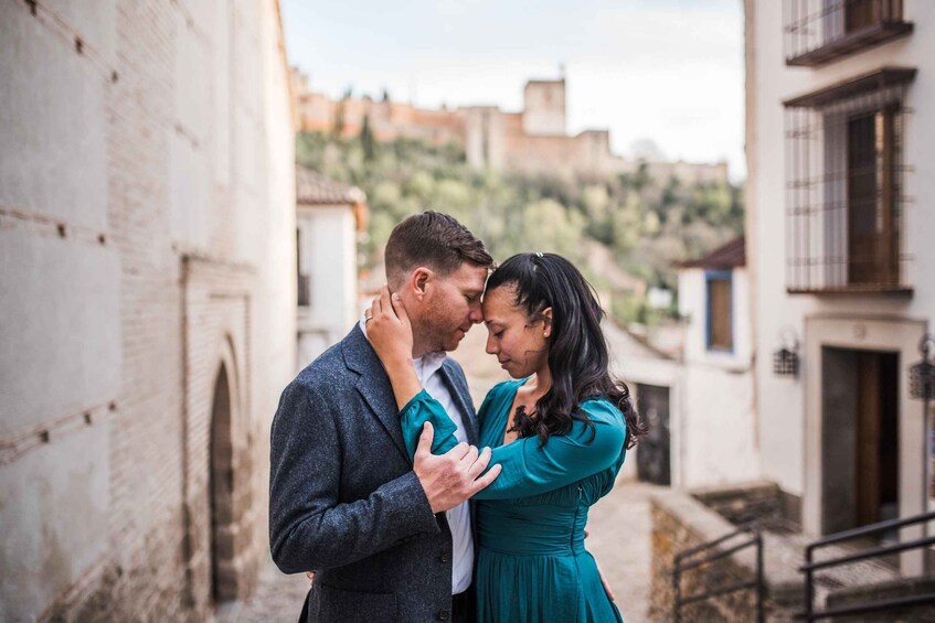A couple embracing in a narrow street with historic buildings and a hill in the background. 1