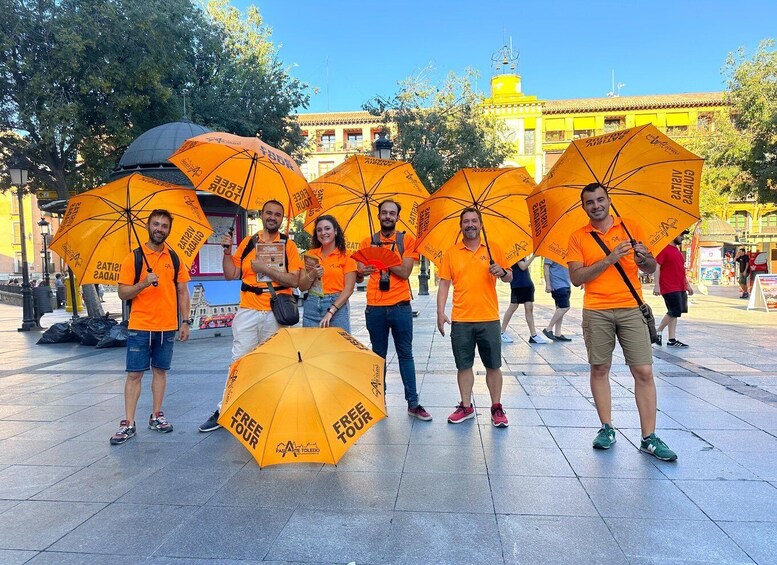 A group of people holding orange umbrellas with "FREE TOUR" text, standing in a plaza with a yellow building in the background. 1