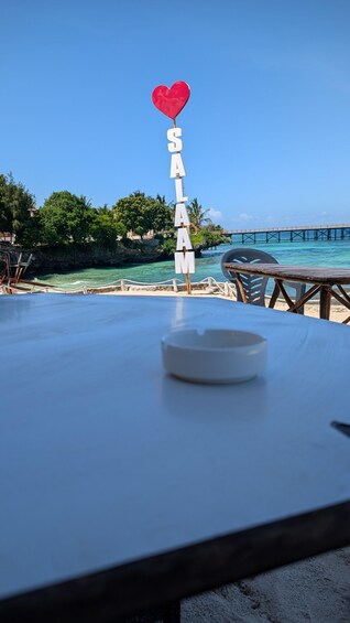 A table with a sign spelling 'SALAMA' and a heart above it, with a beach and pier in the background. 5