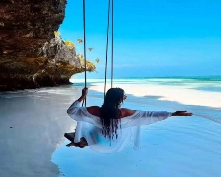 A person swinging on a rope over a beach with clear water and a rocky outcrop. 1