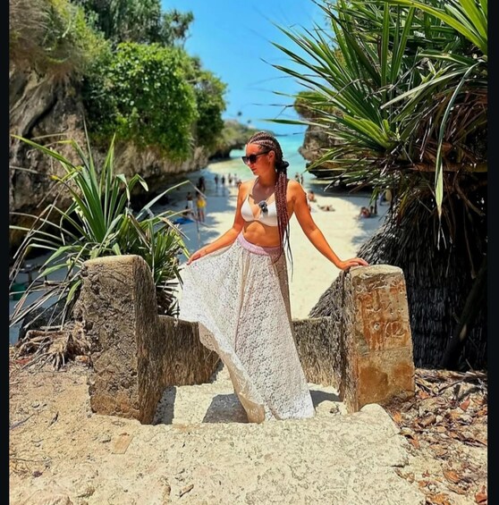 A woman in a bikini and white skirt standing on a stone staircase at a beach with palm trees and a clear blue sky. 4
