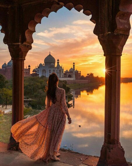 A woman in a flowing dress standing in a pavilion with a view of the Taj Mahal at sunset. 5