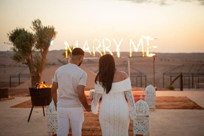 Een stel dat hand in hand staat voor een 'MARRY ME'-bord, met een woestijnlandschap op de achtergrond. 2