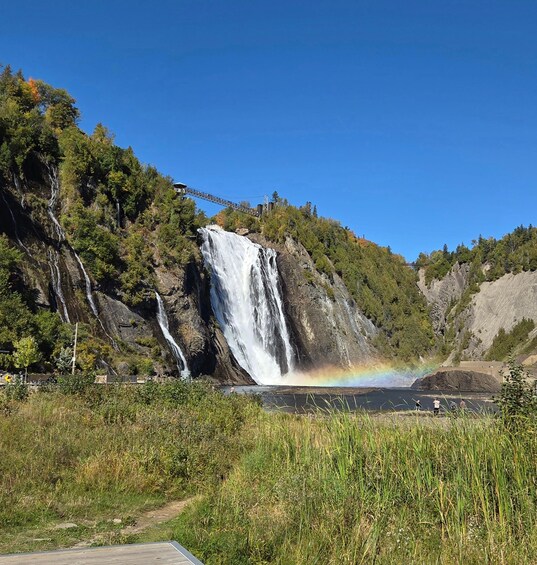 A waterfall with a rainbow, surrounded by greenery and a bridge. 2