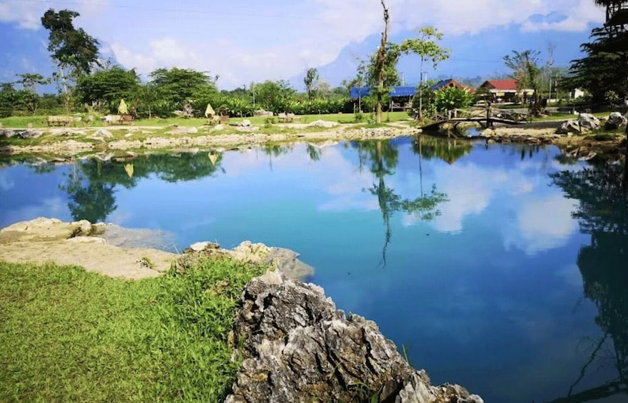 A calm lake with clear reflections, surrounded by greenery and a few buildings in the distance. 2
