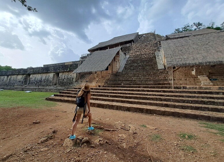 A person standing on a dirt path in front of ancient stone steps leading to a thatched-roof structure. 1