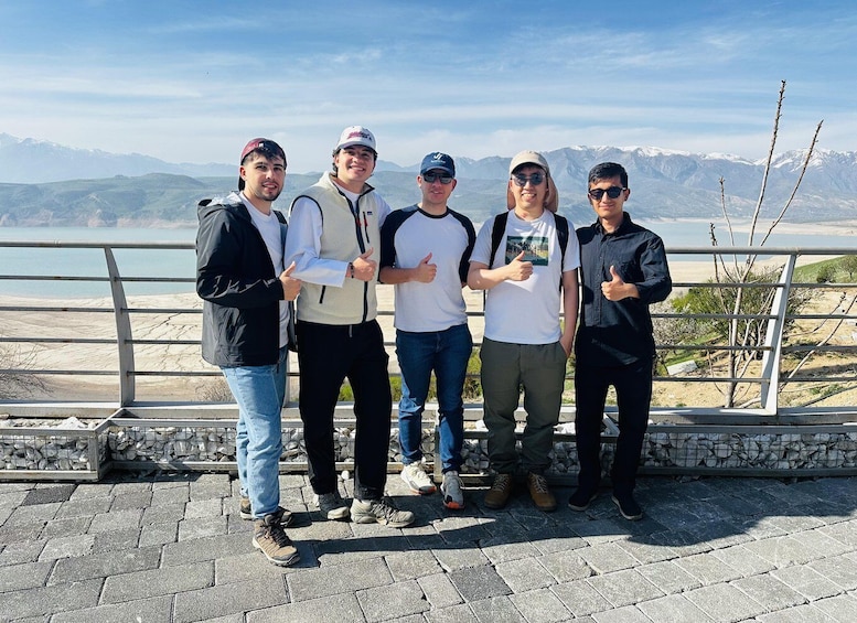 A group of people standing on a paved area with a railing, overlooking a mountainous landscape with a body of water in the distance. 1