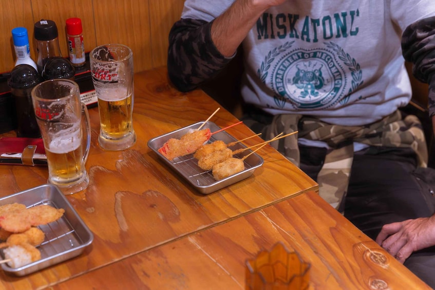 A person sitting at a wooden table with a tray of skewered food, a glass of beer, and condiments. 3