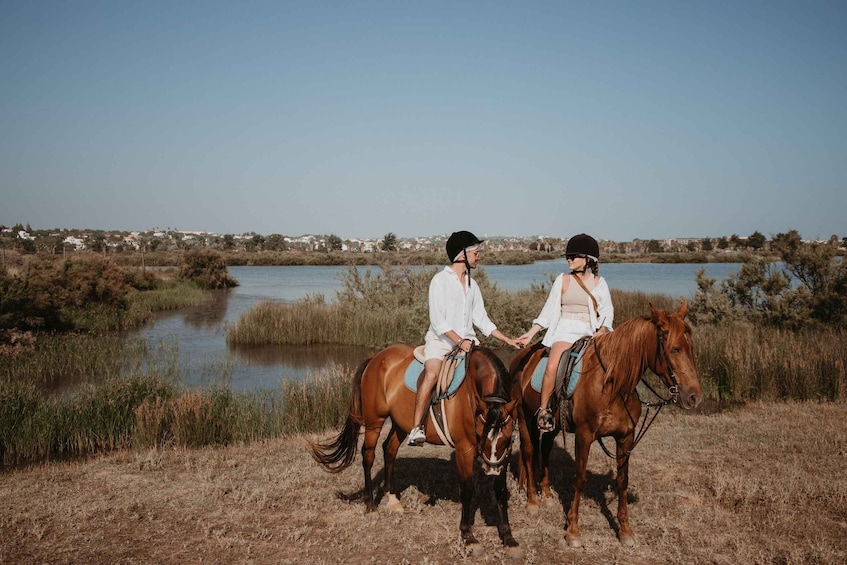 Two people riding horses in a field with a lake and houses in the background. 5