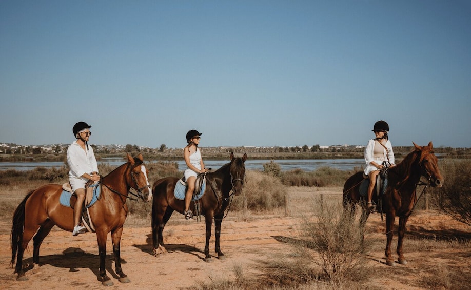 Three people riding horses in a natural landscape with a body of water and distant buildings in the background. 1